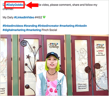 Toto je snímek obrazovky, který ilustruje, jak Goldie Chan používá hashtagy v textu svých videopříspěvků na LinkedIn. Červené popisky odkazují na hashtag #DailyGoldie v textu, který je jedinečný pro její příspěvky ve videu a pomáhá jí sledovat sdílení. Tento příspěvek také obsahuje další relevantní hashtagy, které lidem pomáhají najít její video, včetně #LinkedInVideo. Ve videoobrazu stojí Goldie před některými dveřmi na displeji World of Disney. Je to asijská žena se zelenými vlasy. Měla na sobě černou čepici LinkedIn, černý náhrdelník, růžovou košili s macaronovým potiskem a modrobílou bundu.